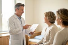 A male doctor in a white lab coat explains medical results to an elderly female patient and a family member in a bright, professional clinic setting. The scene features soft, neutral colors and natural light, capturing a moment of healthcare consultation and patient communication.
