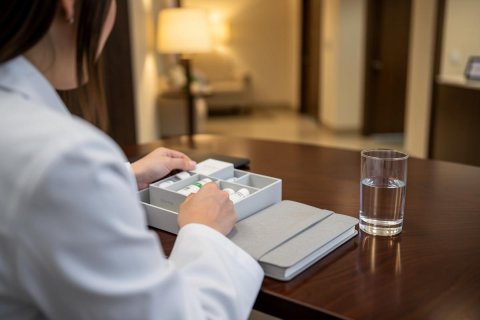 A person in a white lab coat organizes a weekly pill organizer on a dark wooden desk. A glass of water and a notebook sit nearby in a warm, softly lit indoor setting. This scene illustrates medication management, healthcare routines, and wellness in a home or clinical environment.