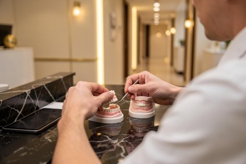 A professional dental technician uses a precision tool to examine detailed dental models on a black marble countertop. The scene is set in a modern, brightly lit dental clinic with a warm, blurred background of a clean hallway. This image highlights dental prosthodontics, laboratory work, and professional oral healthcare in a high-end clinical environment.