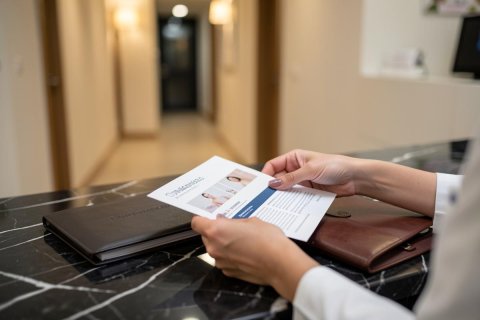 A professional person's hands hold a white printed document over a sleek black marble countertop. Beside the document sit a brown leather portfolio and a black folder. The background features a blurred, brightly lit hallway in a modern office or medical clinic, creating a clean and professional atmosphere.