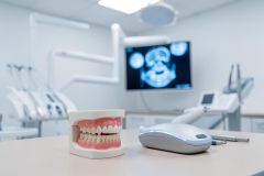 A detailed dental model of teeth and gums sits on a clean surface in a bright, professional dental clinic. The background features blurred dental equipment and a monitor displaying a tooth X-ray, emphasizing oral healthcare. The clinical environment uses a bright white and blue color palette, perfect for dental care and dentistry-related themes.