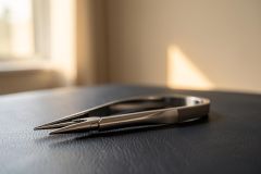 A close-up macro shot of sleek silver metal tweezers resting on a dark, textured leather surface. The background features soft, warm sunlight streaming through a window, creating a beautiful bokeh effect in neutral tones. This minimalist image highlights the precision and clean design of the tool in a serene indoor setting.