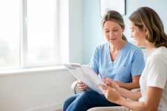 A female nurse in blue scrubs reviews medical documents on a clipboard with a patient in a bright, sunlit clinical setting. This professional healthcare consultation occurs in a modern medical office, highlighting patient care, communication, and medical documentation.
