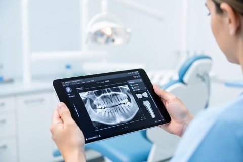 A dental professional holds a digital tablet showing a detailed panoramic dental X-ray in a bright, modern dental clinic. The background features a blue dental chair and clinical white equipment, emphasizing professional oral healthcare and digital dentistry technology. The clean, bright atmosphere highlights advanced medical diagnostics.