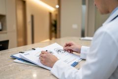 A medical professional wearing a white lab coat works on a clipboard with a detailed anatomical drawing of human eyes. Set in a bright, modern medical clinic with a marble countertop and neutral colors, the scene depicts an ophthalmology consultation or eye examination preparation.