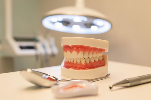 A professional dental clinic setting featuring a realistic model of human teeth and gums on a white surface. Silver dental instruments, including a dental mirror and handpiece, are placed in the foreground. A bright, circular dental examination lamp illuminates the scene in the background, emphasizing oral healthcare and dentistry.