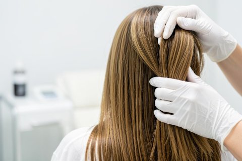 A professional in white medical gloves performs a hair health examination on a person with long, straight light brown hair. Set in a bright, clean clinical environment, the image captures a scalp consultation or dermatological checkup. The focus is on the hair texture and the gloved hands against a soft, neutral background.