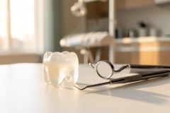 A clear dental aligner and professional dental instruments, including a mouth mirror and probe, rest on a white surface in a brightly lit dental clinic. The warm sunlight and blurred dental equipment in the background create a clean, professional atmosphere for orthodontic and dental procedures.